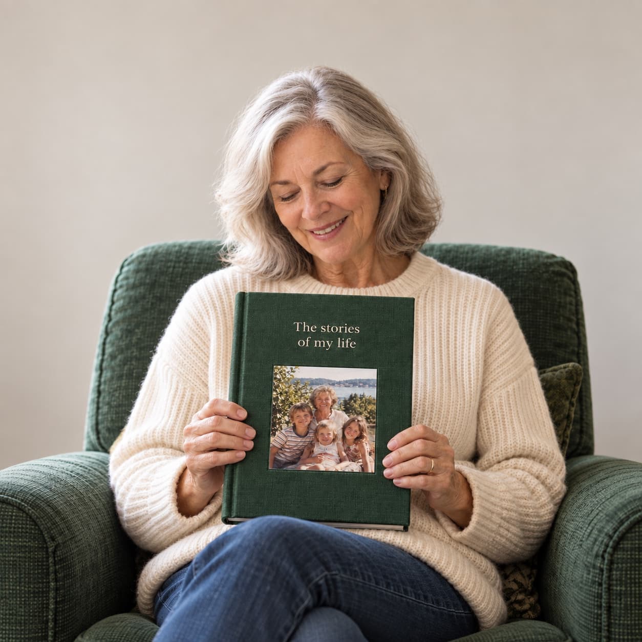 A woman in an armchair smiling down at her finished book, a green linen hardcover titled The stories of my life